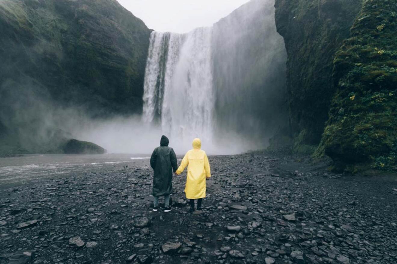 Two people wearing raincoats, standing hand in hand, face a large waterfall surrounded by rocky cliffs.