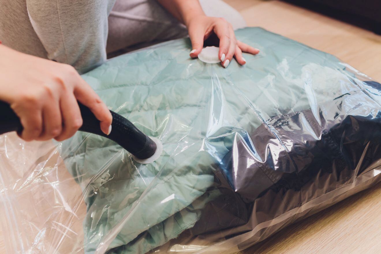 A person uses a vacuum cleaner to compress a green puffer jacket inside a clear vacuum storage bag on the floor.