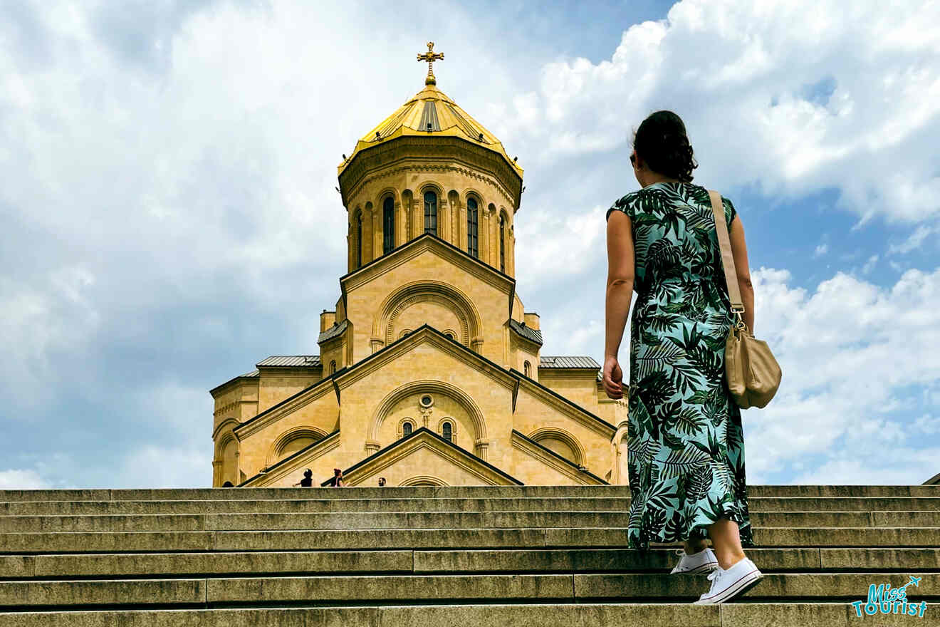 Darija Stojanovic, la autora de las publicaciones de misstourist, con un vestido verde y zapatillas blancas, sube unos escalones de piedra que conducen a una gran catedral ornamentada con una cúpula dorada bajo un cielo parcialmente nublado.