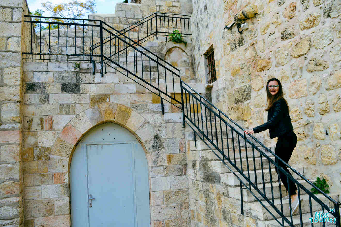 Yulia, la fundadora de este sitio web, con una chaqueta de cuero negra, se encuentra en una escalera de piedra junto a un edificio de piedra con una puerta arqueada cerrada.