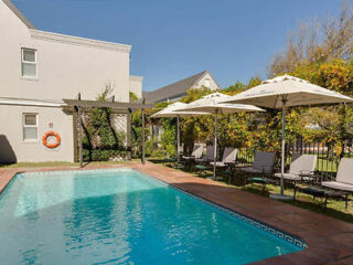 Outdoor swimming pool with lounge chairs and umbrellas on the side, surrounded by greenery and a two-story building in the background under a clear blue sky.