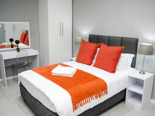 A neatly made bed with gray headboard, orange pillows, and an orange throw blanket in a room with white walls, a white wardrobe, a small desk with a mirror, a chair, and bedside tables with lamps.