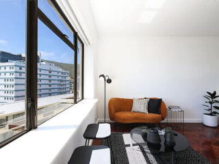 A minimalist living room with a large window, an orange sofa, a floor lamp, and a black table with decorative items. A potted plant is in the corner, and a cityscape is visible outside the window.