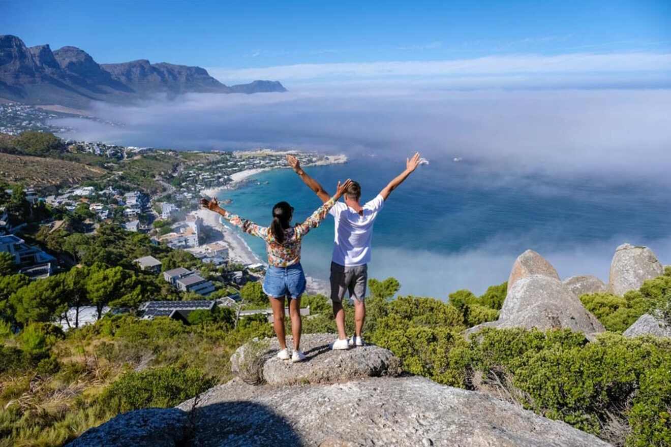 Two people stand on a rocky outcrop with arms raised, overlooking a coastal town, mountains, and a foggy seascape.