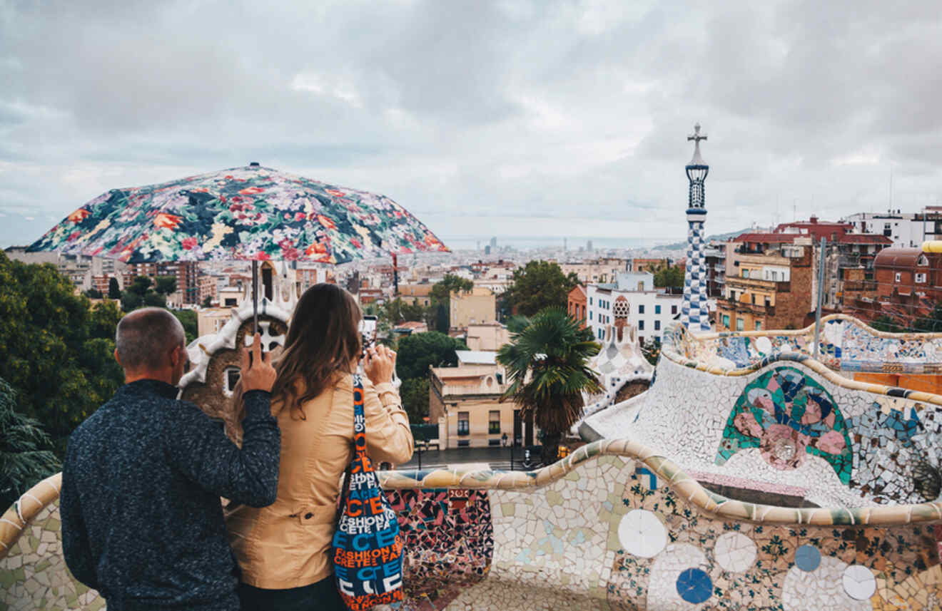 Two people standing in Park Güell, Barcelona, under a colorful umbrella with a scenic view of the city in the background, indicating rainy weather