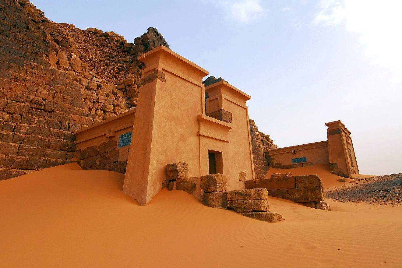 Ancient pyramid structures partially buried in sand, with weathered walls and stone steps. Clear blue sky in the background.