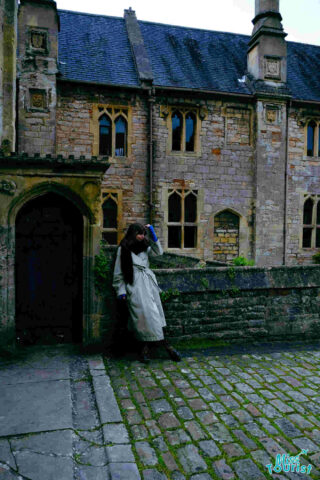 The writer of the post in a trench coat leaning against a historic stone building with arched windows and cobblestone path