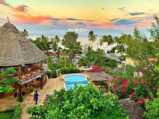 A tropical resort with a thatched-roof building, a swimming pool, lush greenery, and colorful flowers at sunset. Palm trees and an ocean are visible in the background.