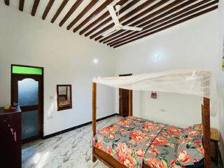 A bedroom with a floral-patterned double bed, wooden four-poster frame with a white canopy, marble floor, wooden beamed ceiling, ceiling fan, wall mirror, and two doors.