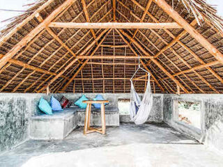 A rustic loft with a thatched roof features a hammock, a wooden table, and stone benches adorned with colorful cushions. The space is open and airy, allowing natural light to filter through.