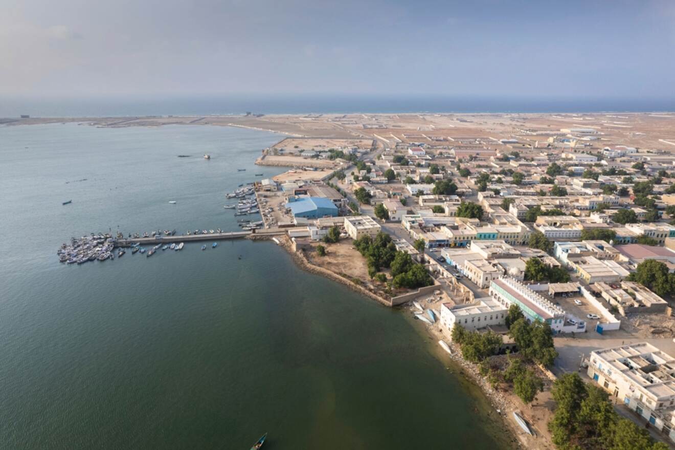 Aerial view of a coastal town with a pier extending into the water, numerous buildings, and arid landscape in the background.