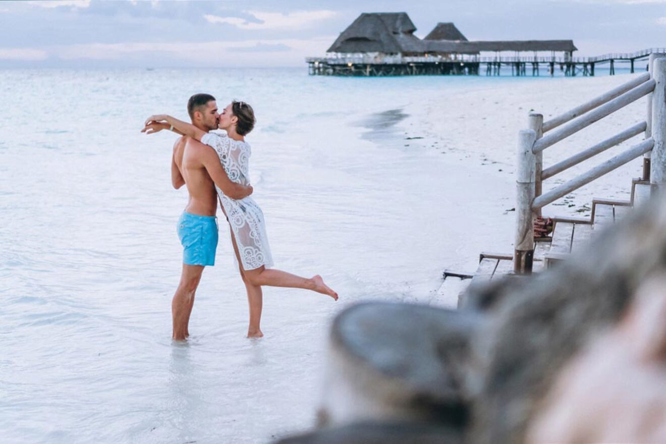 A couple shares a kiss while embracing at the water's edge on a beach. The woman is lifted off the ground, and they are near a wooden pier with thatched-roof structures extending over the water.