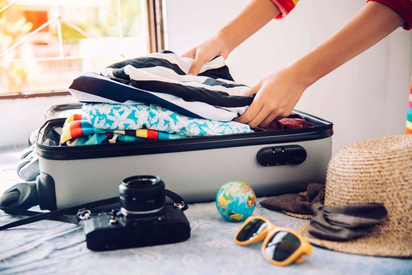 A person packs clothes into a suitcase on a bed, while a camera, small globe, hat, sunglasses, and a watch lie nearby.