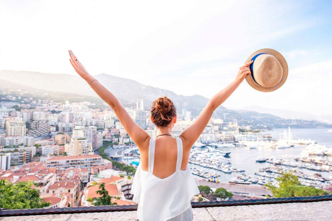 A woman with arms raised and holding a hat looks out over a city with numerous buildings and a marina filled with boats.