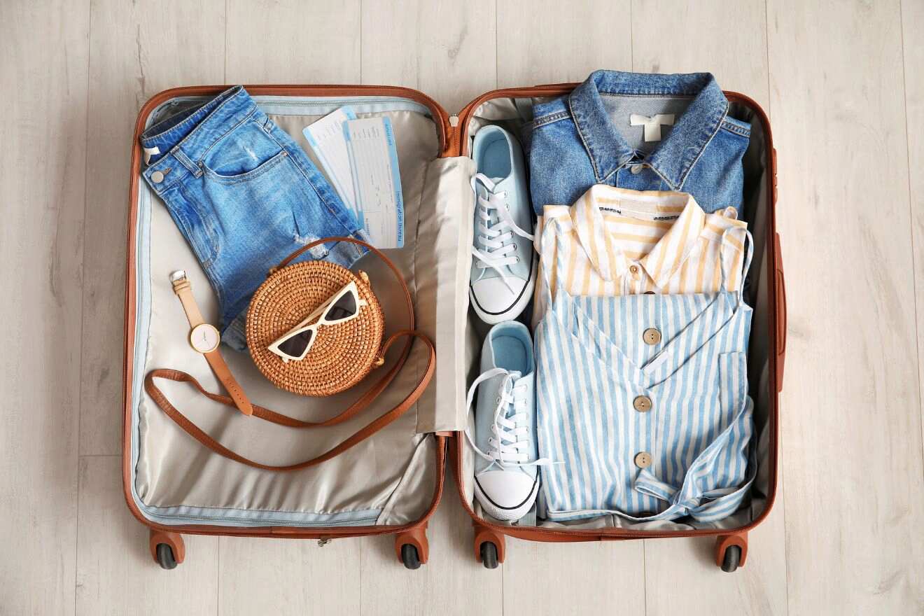 An open suitcase on a wooden floor contains neatly folded clothes, a pair of sneakers, a straw handbag with sunglasses, a wristwatch, and travel documents.
