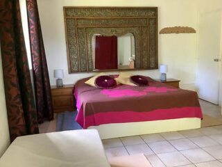 A bedroom with a double bed featuring a brown and pink bedspread, two decorative pillows, bedside tables with lamps, a large ornate mirror above the bed, and tiled flooring.