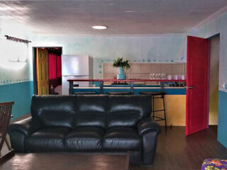 A living area with a black leather sofa, a wooden coffee table, and an open kitchen in the background. The kitchen has a small counter with stools, a refrigerator, and shelves with dishes.