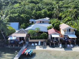 A scenic beachfront property with multiple buildings surrounded by lush greenery, featuring a dock, kayaks, and lounge chairs on the sandy shore.
