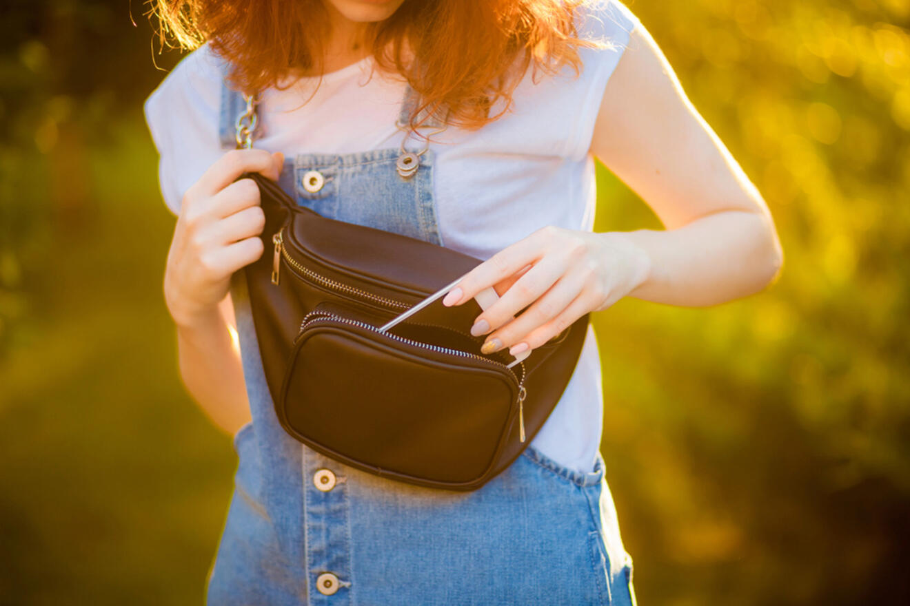 A person with long hair wearing a light blue shirt and denim overalls is opening a black waist bag outdoors in sunlight.