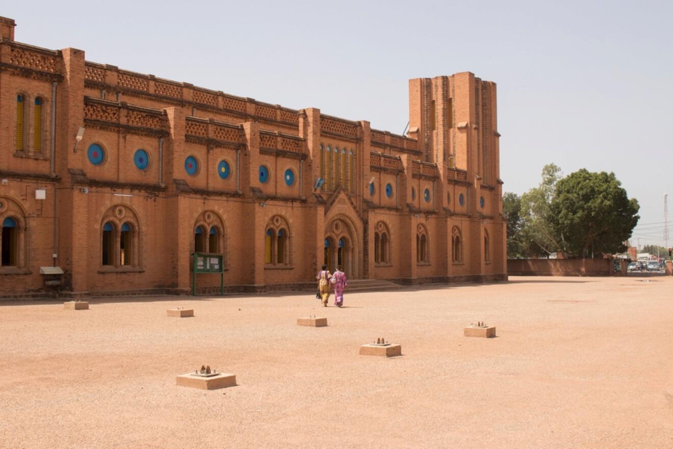 A large, reddish-orange brick building with blue circular details on the windows. Three people walk outside, surrounded by a spacious, sandy courtyard with a few rectangular bases.