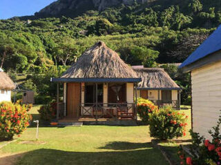 A small house with a thatched roof, surrounded by green bushes and trees, is set against a lush, mountainous backdrop.