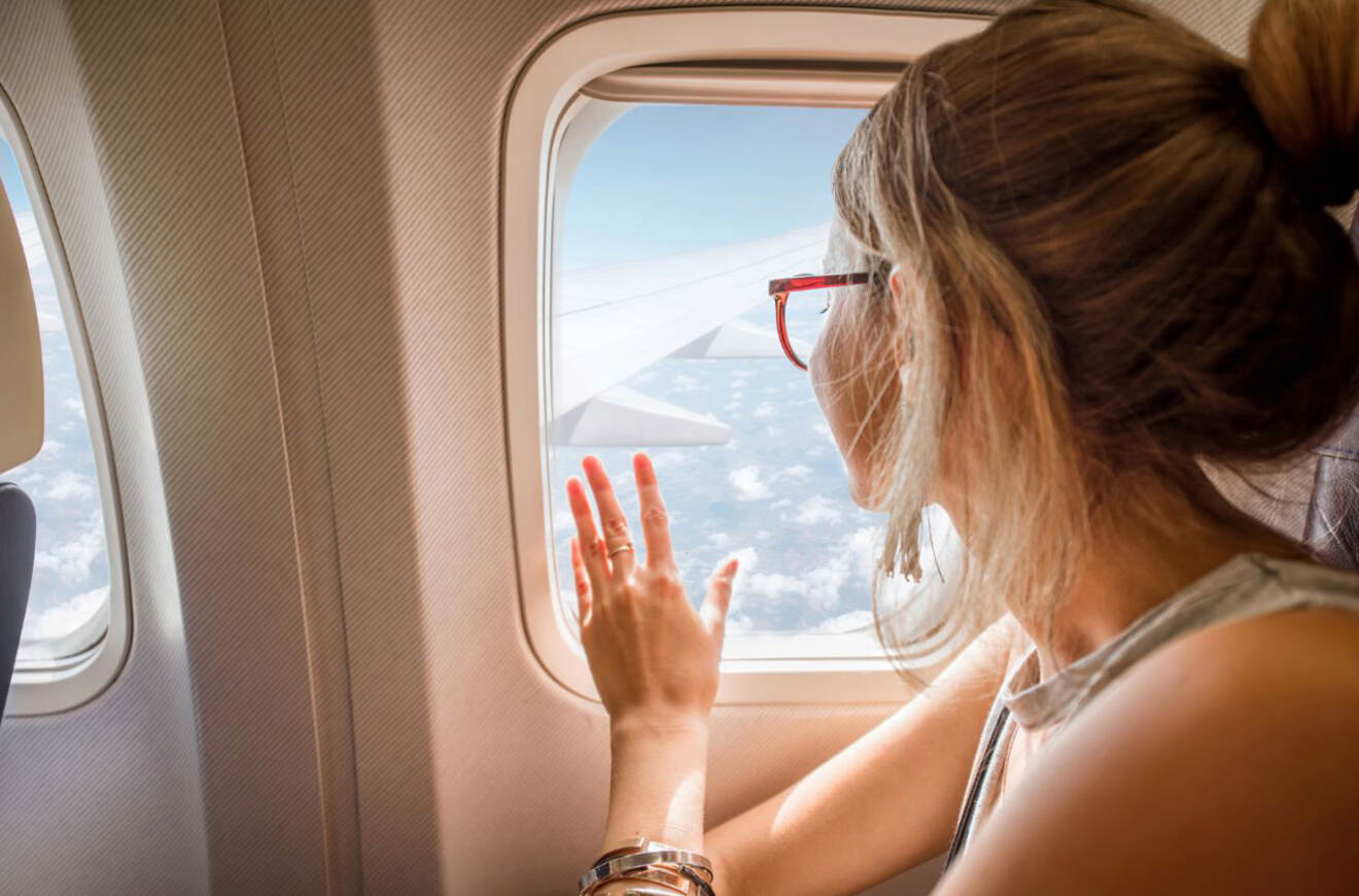 A woman with a bun and glasses looks out the airplane window, with a wing and clouds visible outside.