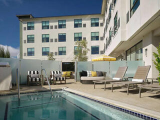 Hotel courtyard with outdoor pool, lounge chairs, and seating area under a yellow-striped umbrella, adjacent to a multi-story building with multiple windows.