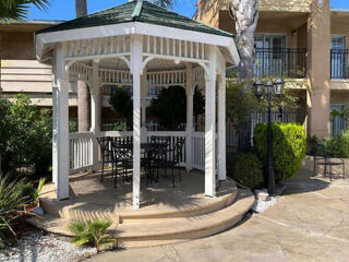 A white wooden gazebo with a green roof in a courtyard, surrounded by greenery. Inside, there is a black metal table with chairs. Two-story buildings are visible in the background.