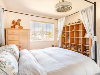 A bedroom with a four-poster bed featuring floral pillows, a wooden dresser topped with a horse figurine, a large window, and a wooden shelving unit filled with books and decorations.