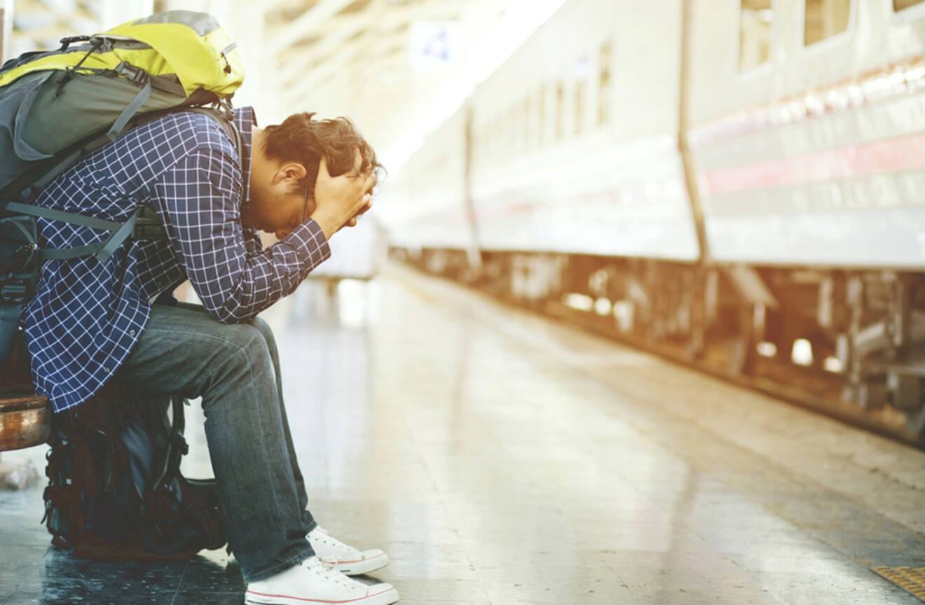 Traveler sitting at a train station with head in hands, looking stressed or worried, with a backpack beside them and a train in the background.