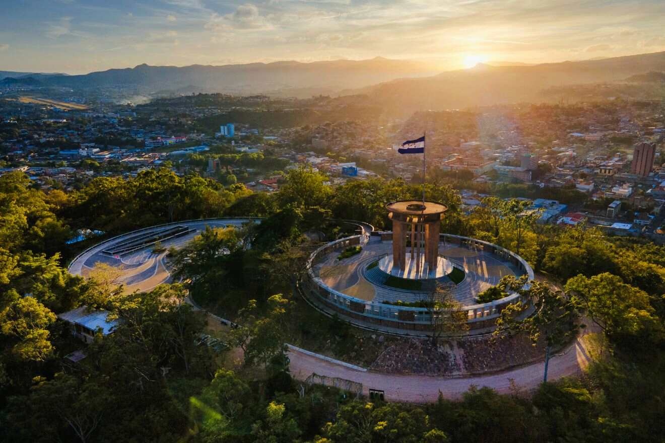 Aerial view of a circular structure with a tall central tower surrounded by greenery at sunset. A flag flies on the central tower. The city landscape extends to the horizon with mountains in the background.