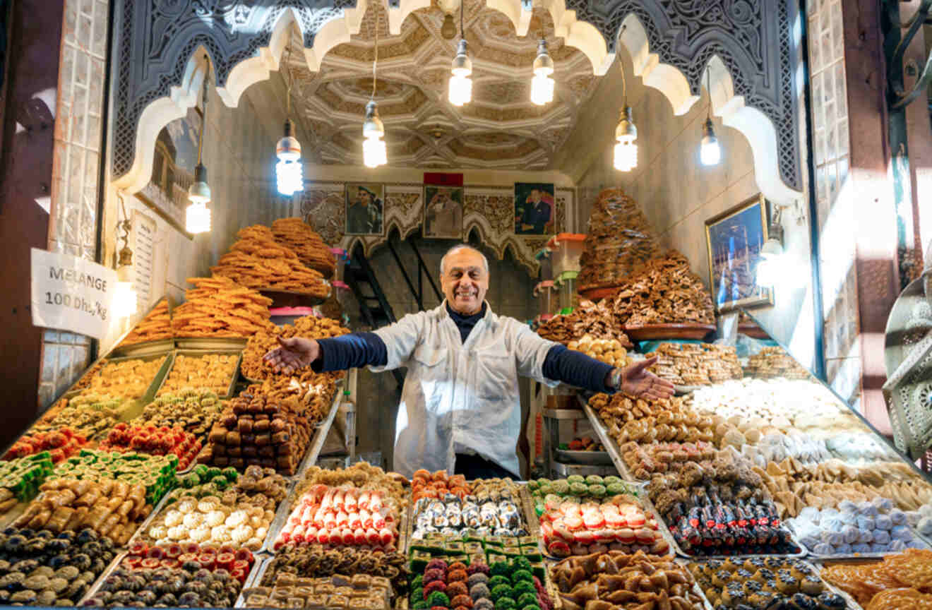 Man with open arms behind a colorful display of sweets at a market in Marrakech, Morocco.