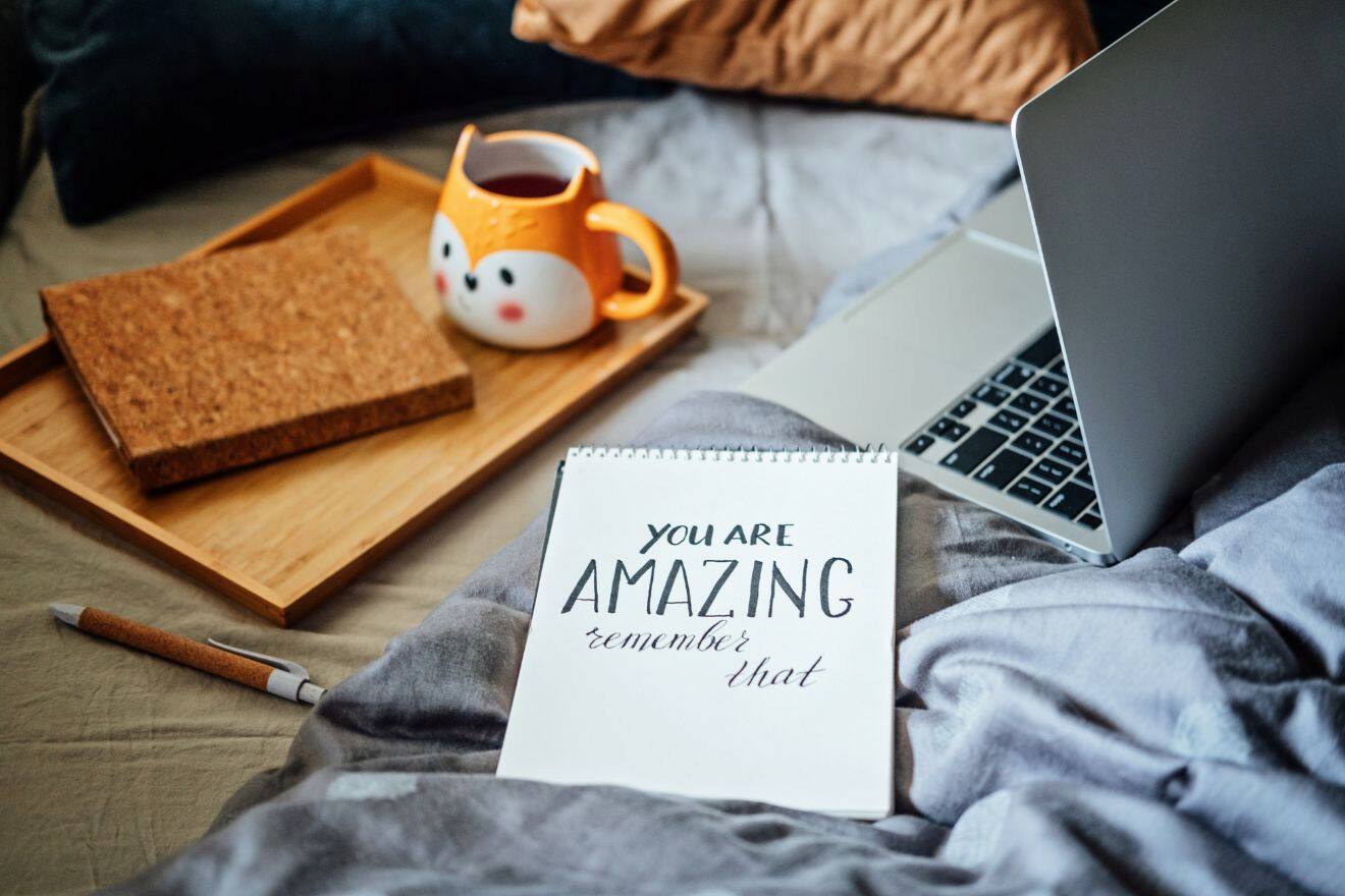 A cozy bed with a notebook that says "You are amazing, remember that," a laptop, a pen, a tray with a cat-themed mug of tea, and a notebook.