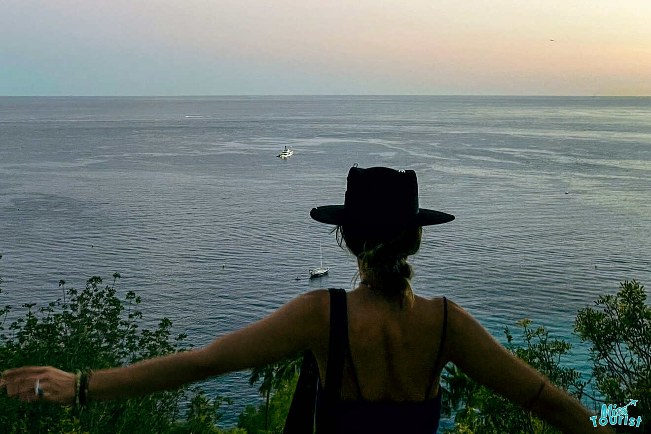 Victoria Lupascu, autora de las publicaciones o misstourist, con sombrero negro y top oscuro, se encuentra con los brazos extendidos, mirando al océano desde un mirador elevado al atardecer. A lo lejos se ve un barco en el agua.