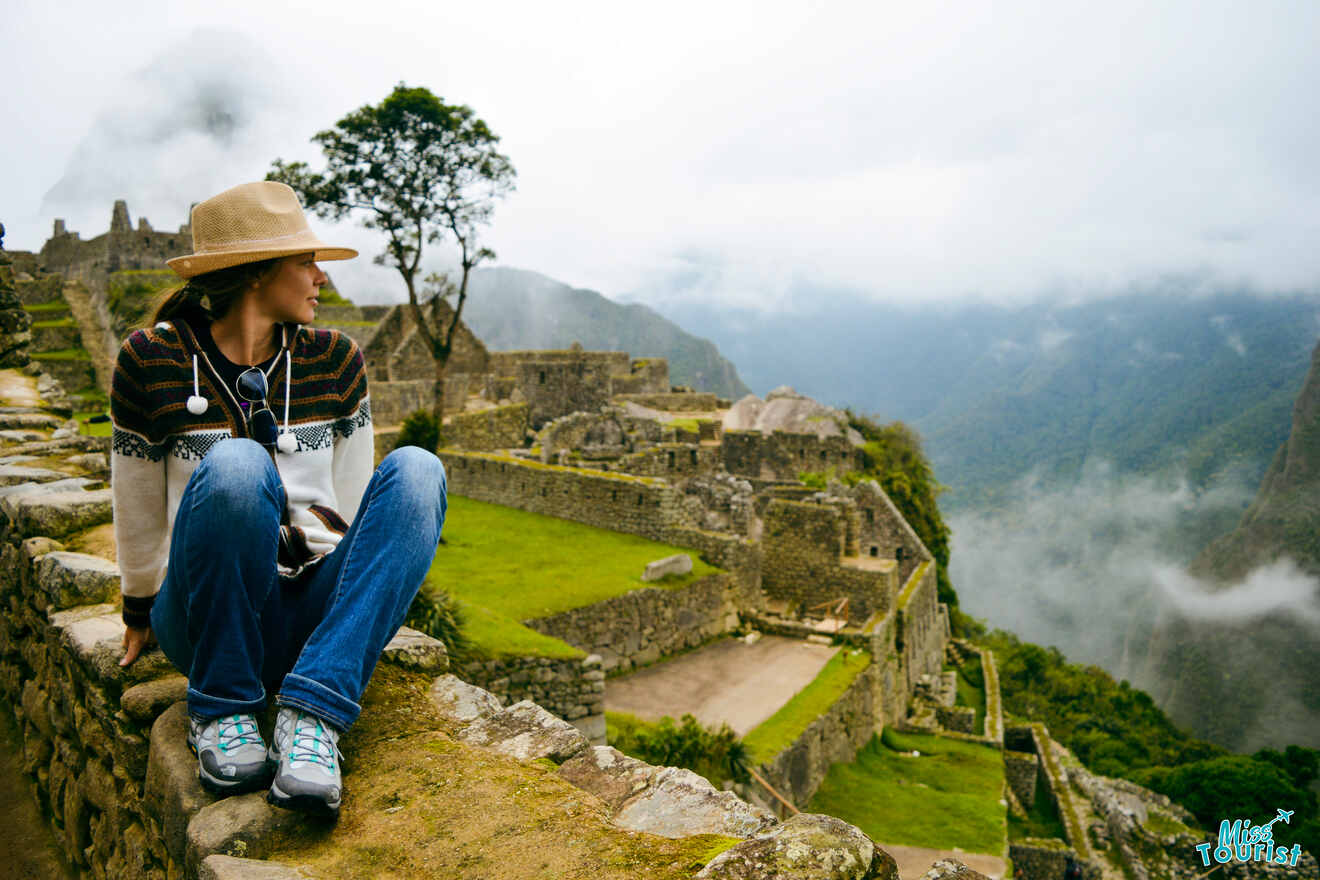 Yulia, la fundadora de este sitio web, vestida con sombrero y atuendo informal, se sienta en una cornisa de piedra con vistas a las ruinas de Machu Picchu con un fondo de montañas brumosas.