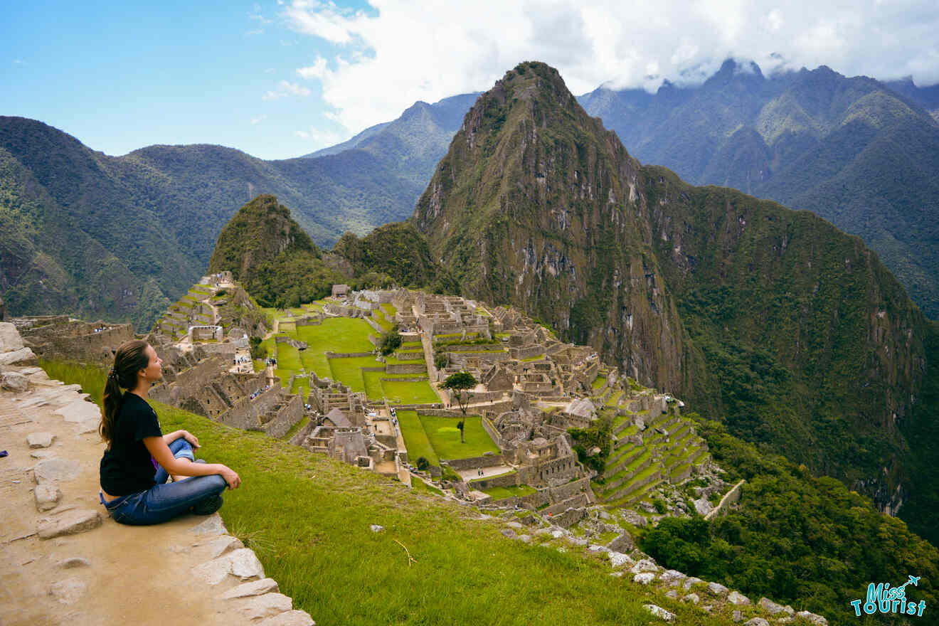 Yulia, la fundadora de este sitio web, se sienta en una cornisa cubierta de hierba con vistas a la antigua ciudad inca de Machu Picchu, rodeada de montañas verdes y un cielo parcialmente nublado.