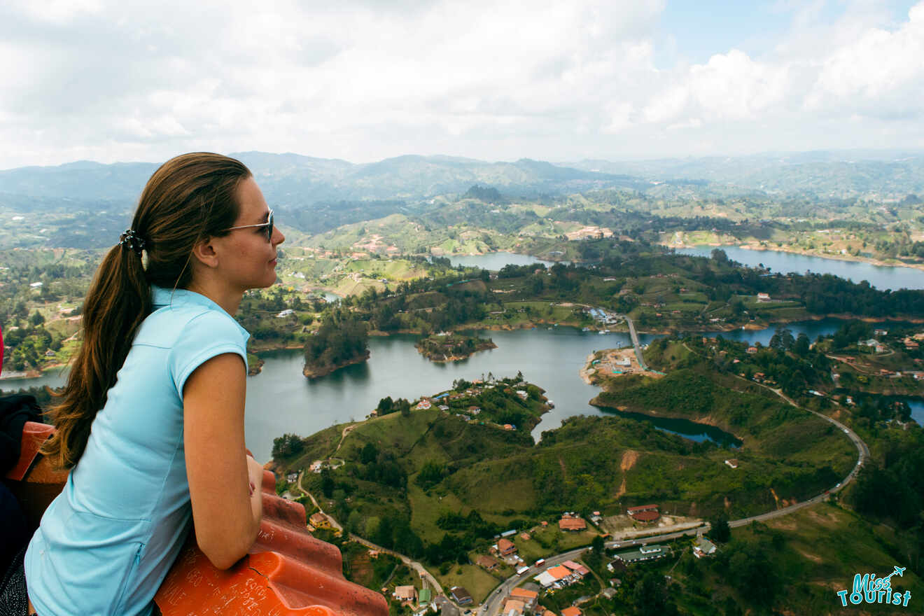 Yulia, la fundadora de este sitio web, con gafas de sol y una camisa azul contempla un paisaje pintoresco con un lago y vegetación, visto desde un punto elevado.