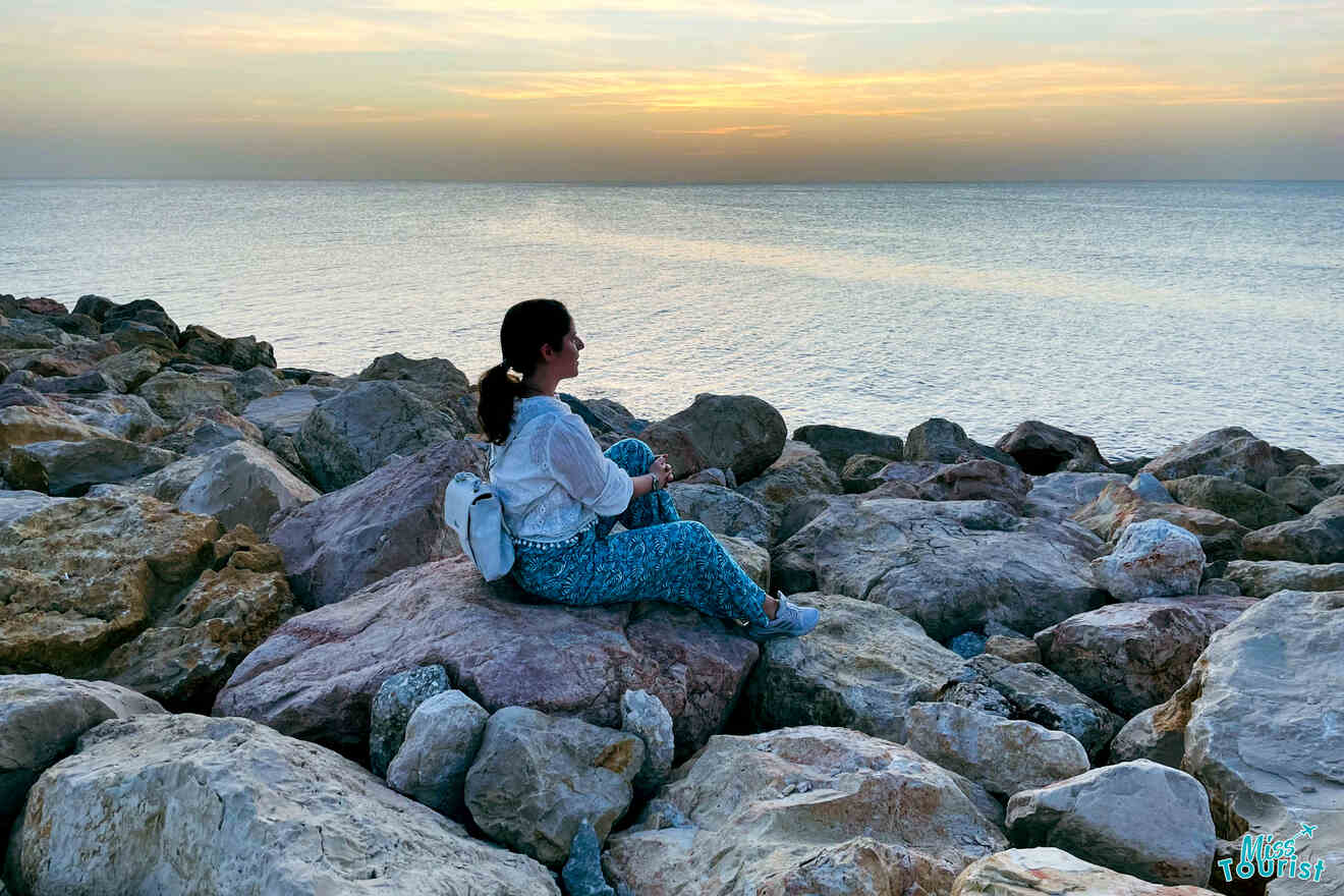 Darija Stojanovic, la autora de las publicaciones o misstourist, vestida con una camisa blanca y pantalones azules y con una mochila blanca, está sentada en las rocas junto al mar, mirando hacia el horizonte durante la puesta de sol.