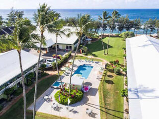 Aerial view of a resort with a swimming pool, green lawn, palm trees, and buildings near a beach with the ocean in the background.