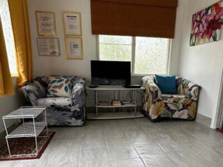 A small living room with two flower-patterned armchairs, a TV on a stand, and framed artwork on the walls. The floor is tiled and a window with a brown blind is in the background.