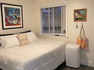 A small bedroom with a neatly made bed, a pillow with "smile" text, wall art, a window with blinds, and a portable air conditioner next to hooks holding a bag.