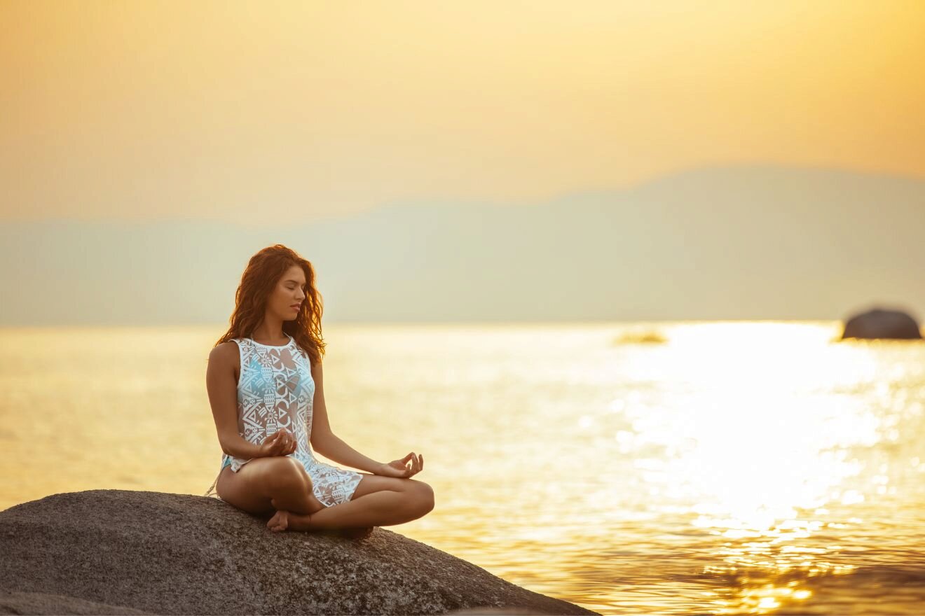 A woman in a white dress sits cross-legged on a rock near a body of water at sunset, meditating with hands resting on knees.
