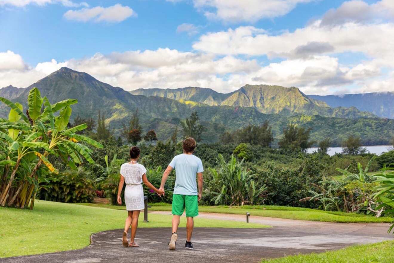 A couple holds hands while walking on a paved path surrounded by greenery, with mountains and a partly cloudy sky in the background.