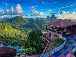 Scenic view of a lush mountainous landscape with a building featuring a red roof and colorful tiles in the foreground. The sky is partly cloudy with clear blue patches.