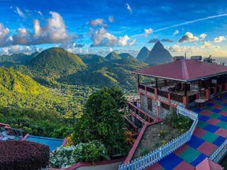Scenic view of a lush mountainous landscape with a building featuring a red roof and colorful tiles in the foreground. The sky is partly cloudy with clear blue patches.