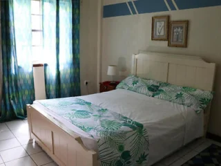 A bedroom with a white bed, green leaf-patterned bedding, two wall pictures above the headboard, a red nightstand with a lamp, and a window with blue and green patterned curtains.