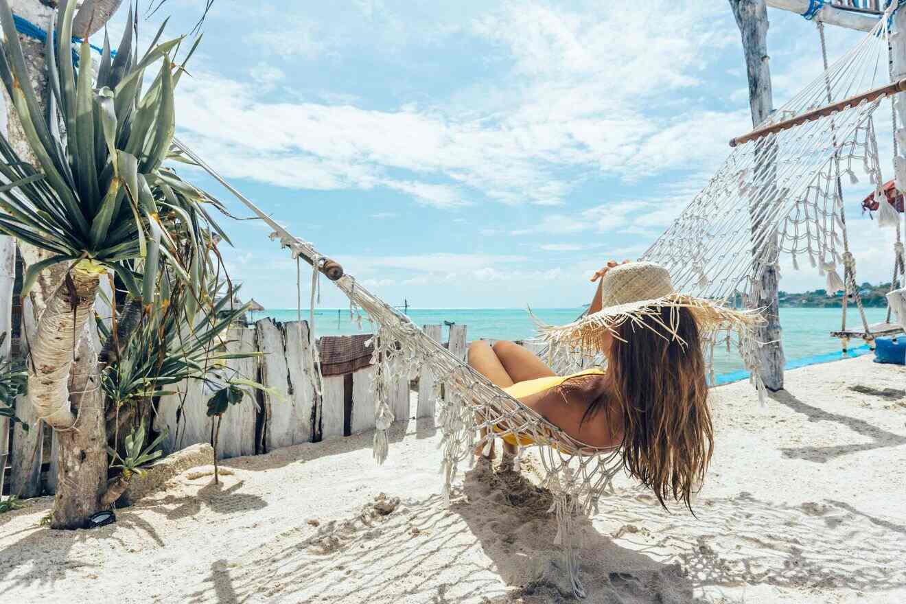 Woman in a straw hat relaxes on a hammock by the beach, overlooking the turquoise sea. Surrounding area includes a wooden fence and tropical plants under a sunny sky.