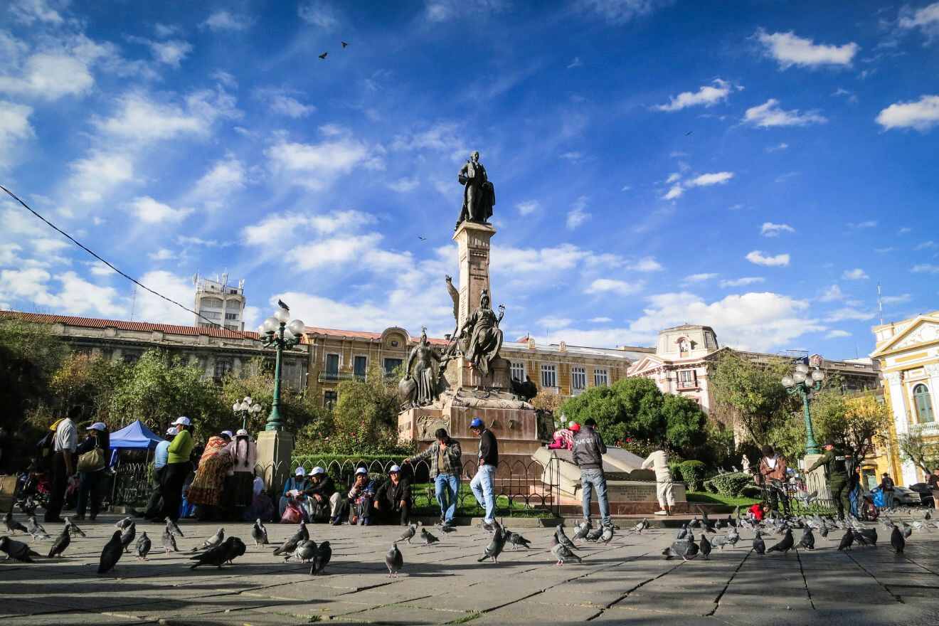 People gathering around a large statue with pigeons on a sunny day in a public square surrounded by historic buildings.