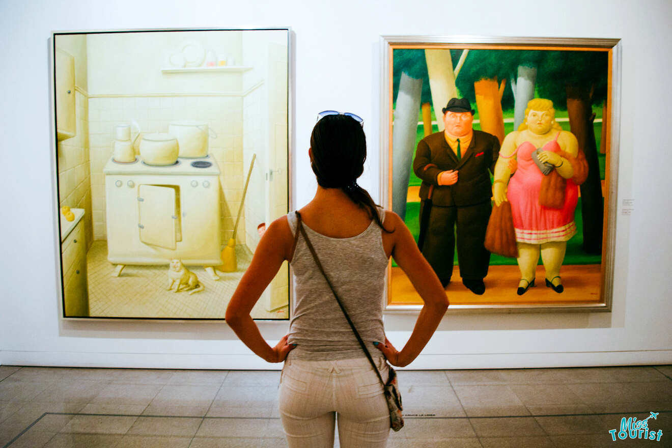 Yulia, la fundadora de este sitio web, se encuentra frente a dos pinturas, una de una escena de cocina y otra de dos figuras vestidas con atuendos formales, en una galería de arte.