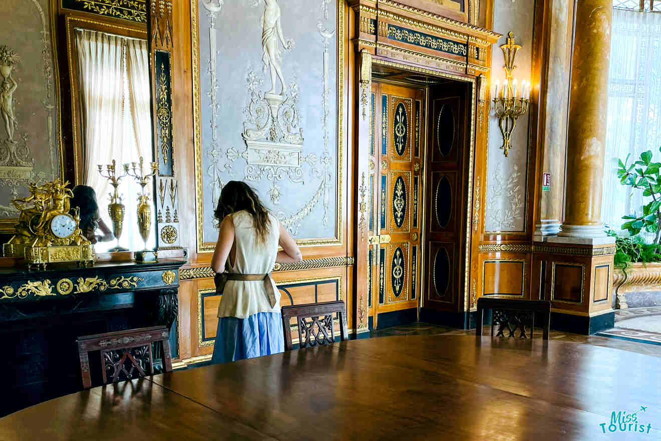 Victoria Lupascu, la autora de las publicaciones de misstourist, con cabello largo, se encuentra frente a una chimenea ornamentada en una habitación ricamente decorada con elaborados diseños de paredes y muebles de madera.